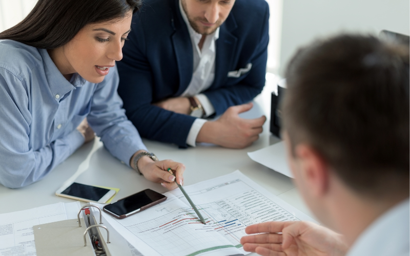 Reunión entre un asesor financiero y dos clientes revisando gráficas, analizando situación financiera.